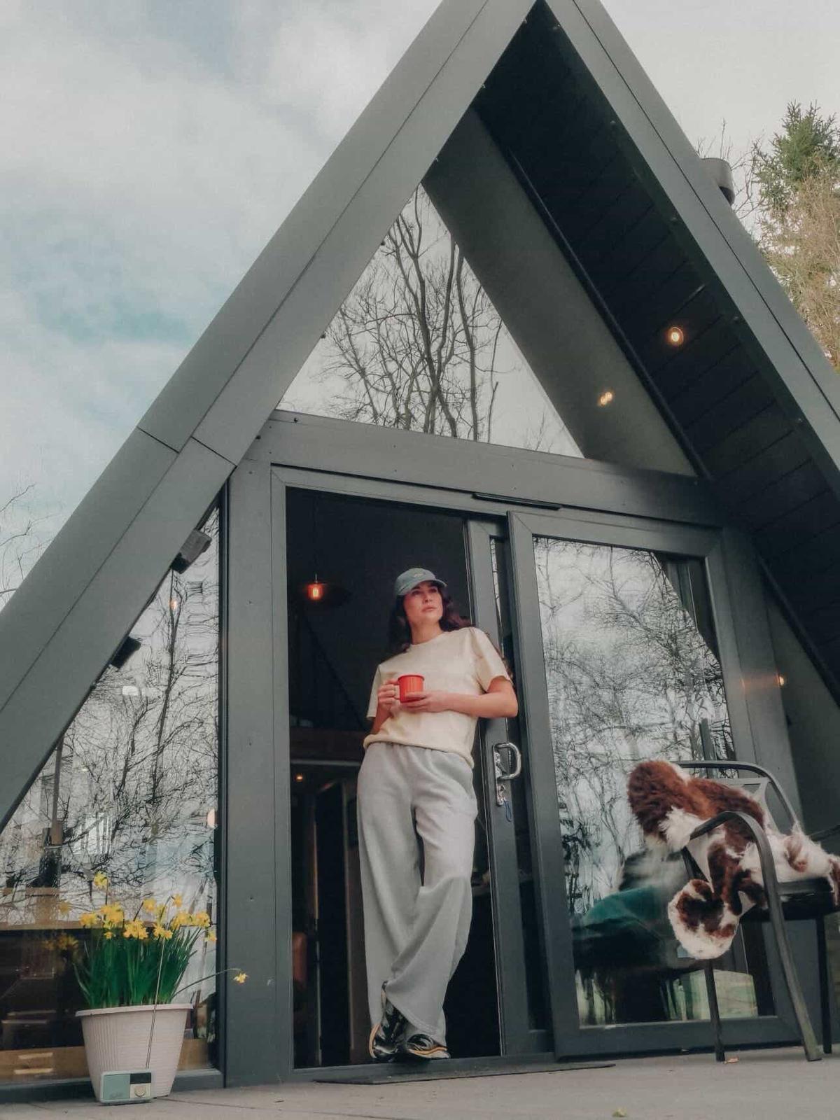Fashion shoot model standing in the doorway of an A-frame cabin holding a red mug